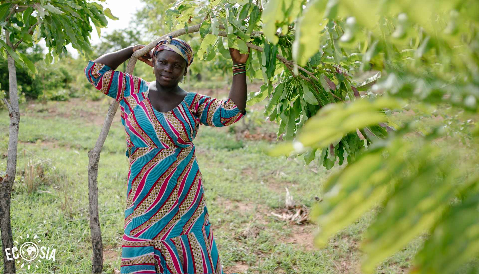 A women, Makada, in a brightly coloured dress under a tree, photo credit Ecosia.