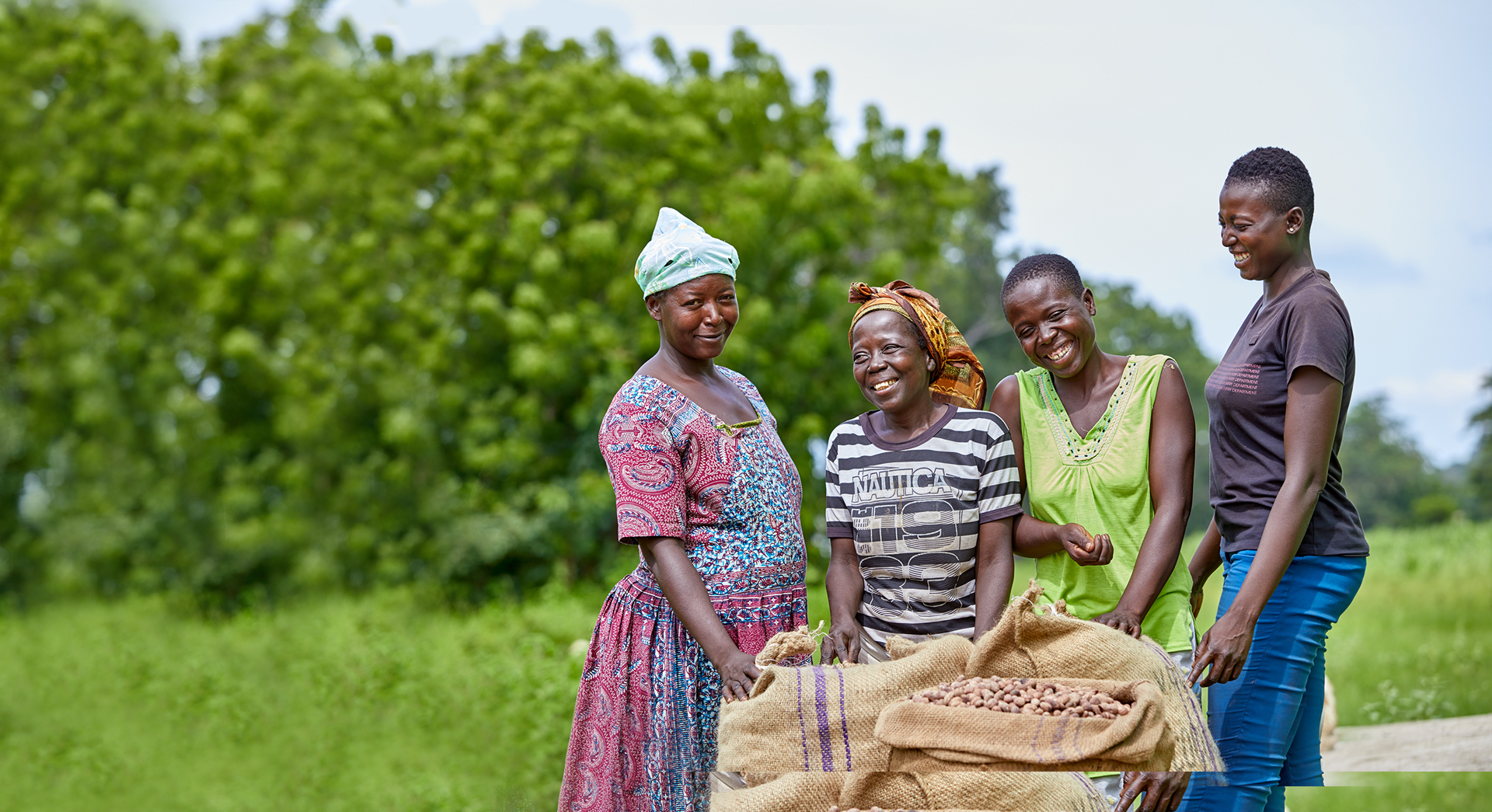 A group of women that are part of a shea enterprise group standing around bags of shea nuts they have collected and smiling and laughing.