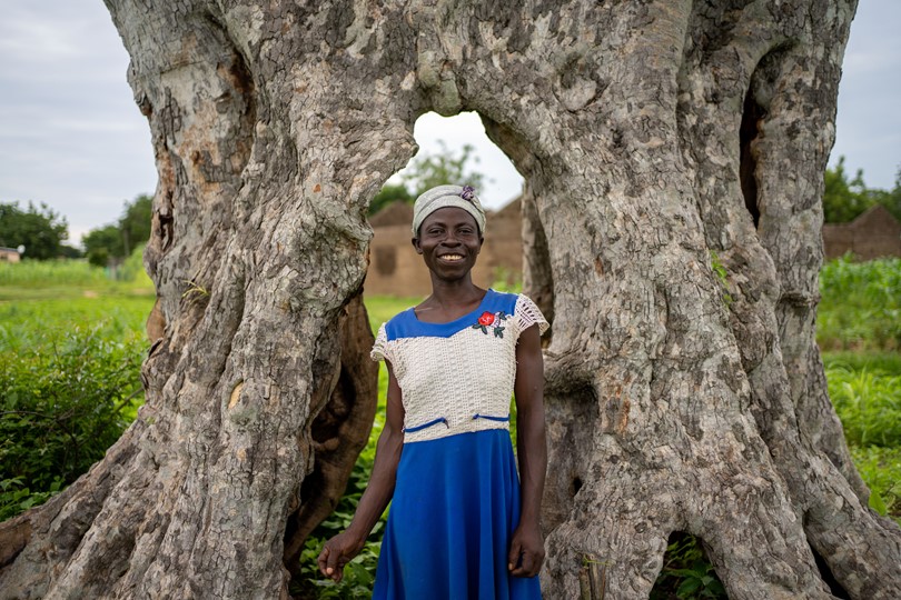 A woman in Ghana smiling, standing in front of a giant tree trunk