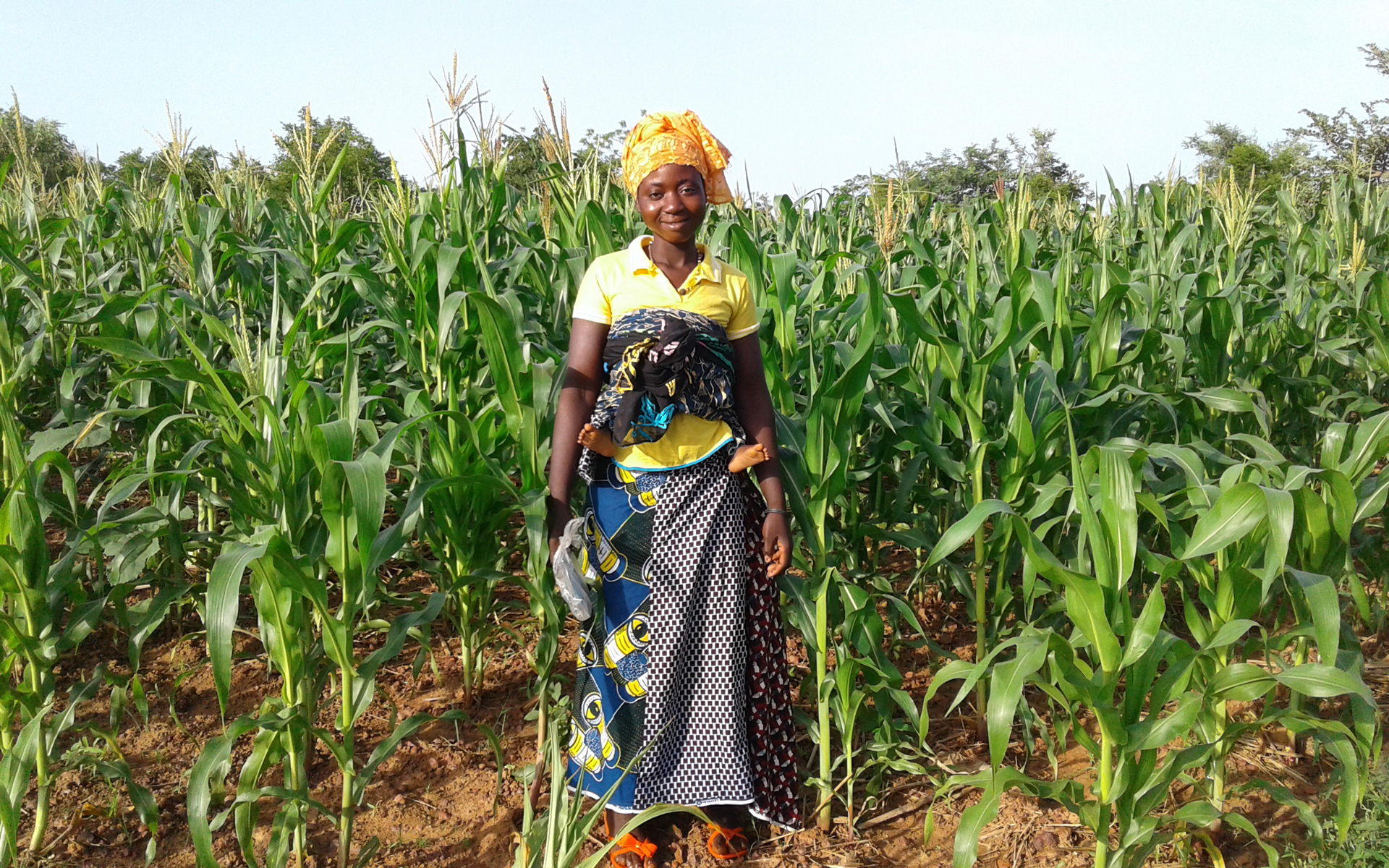 A women standing and smiling in her farm where she is growing crops for food and to sell.
