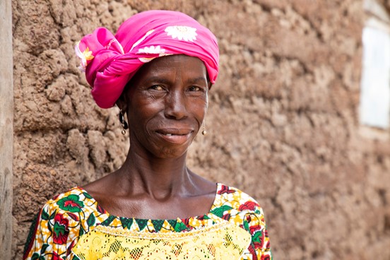 Portrait of Habi stood outside her home in south-western Mali. She is wearing a bright pink scarf on her head, and a yellow dress with green and red flowers on it. She is smiling at the camera.