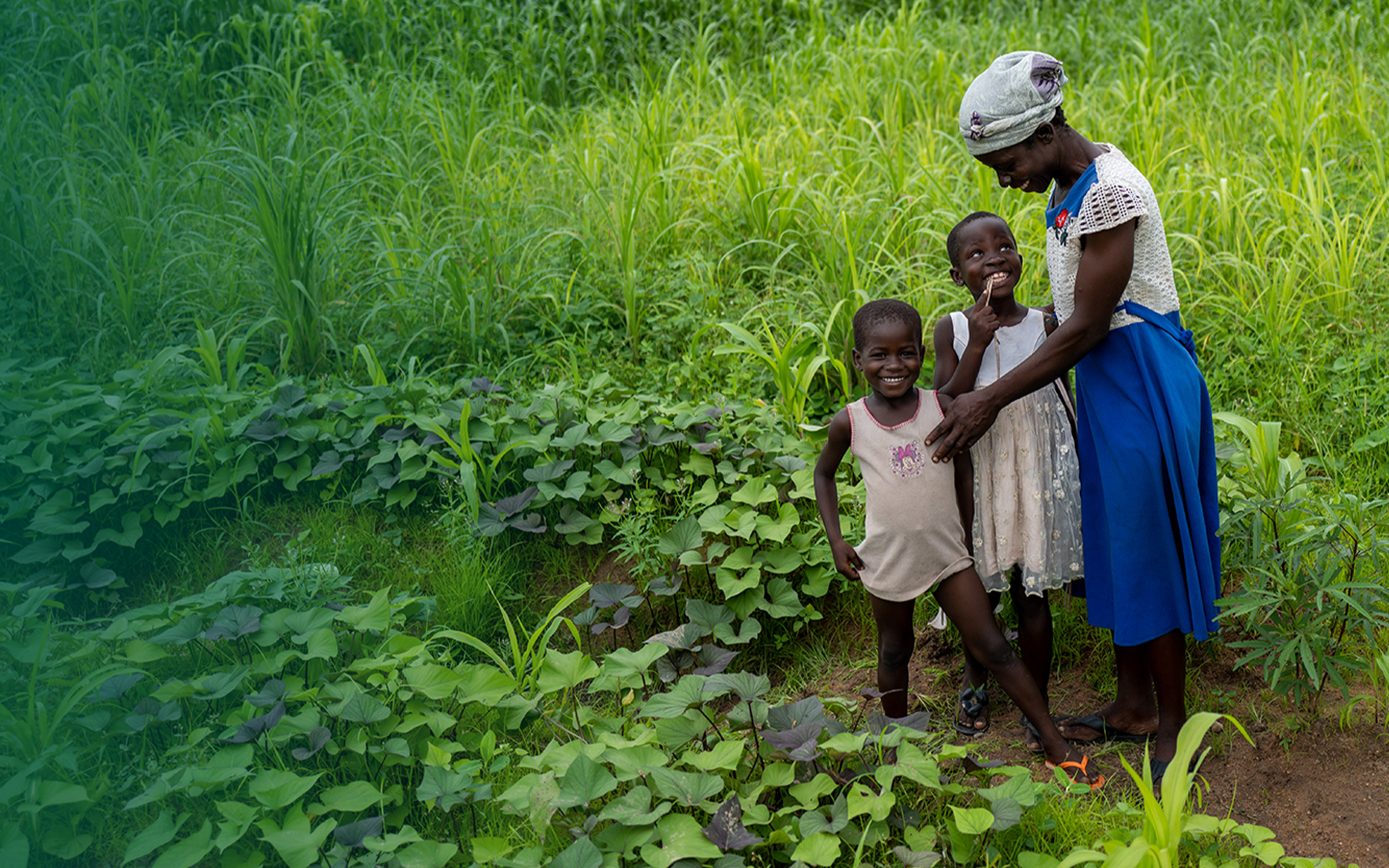 Abba Achana standing and smiling with her two children in a field, © 2021 Tree Aid.