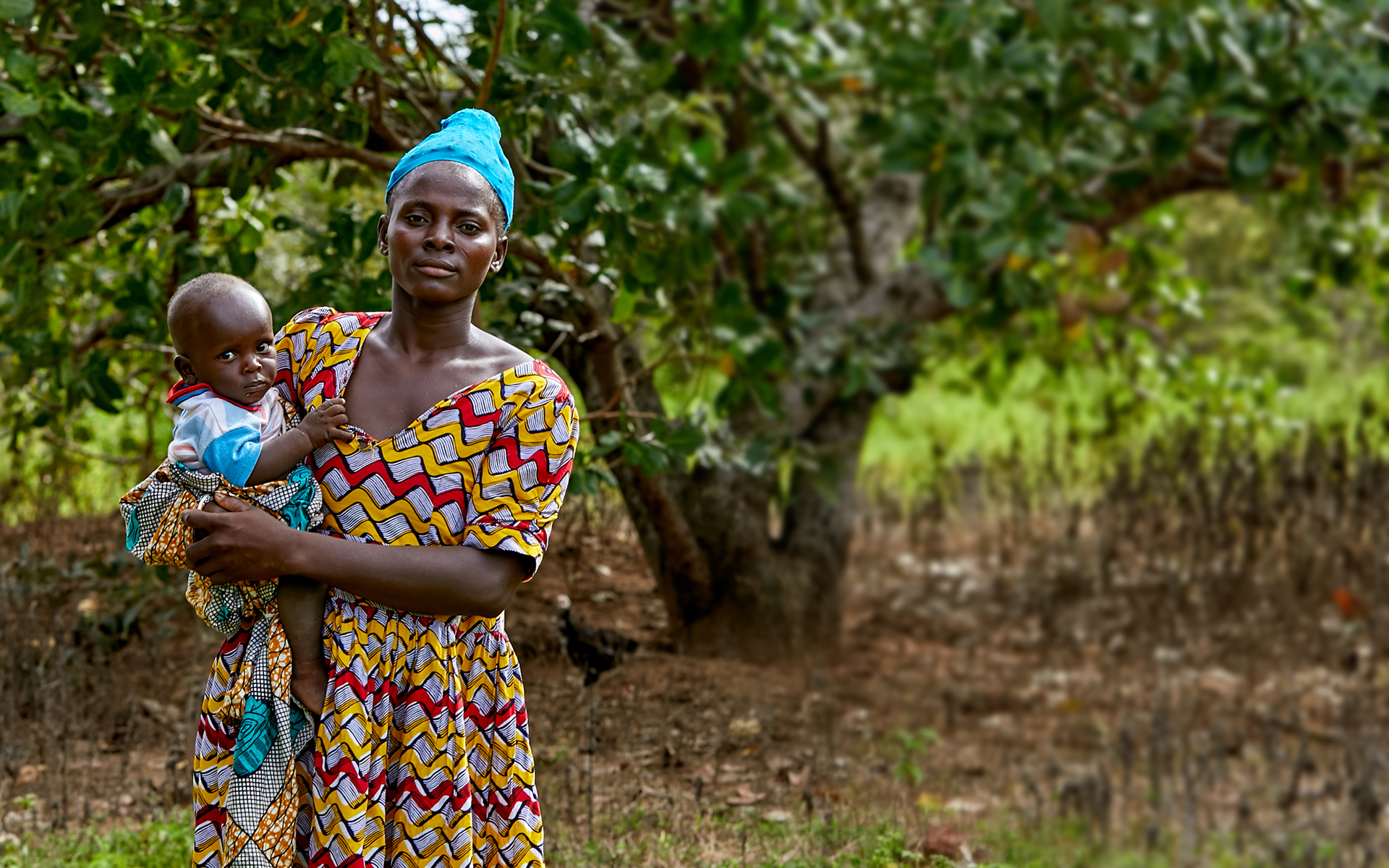 Gifty is holding her baby in front of a tree. She is being supported by a Tree Aid project to earn an income from cashew trees.