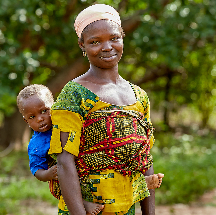 Elizabeth, a mother in Ghana, is standing in front of a cashew tree smiling with her son strapped to her back.