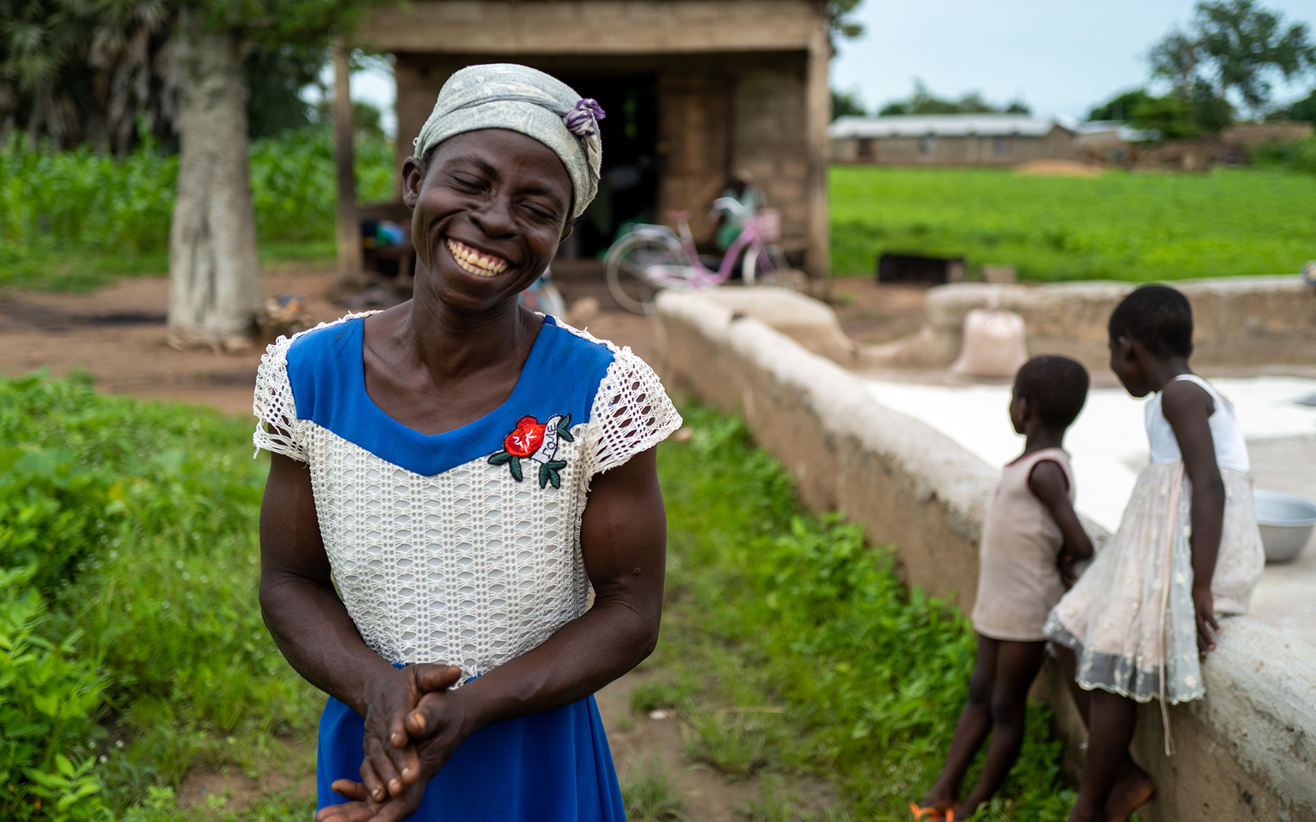 Abba Achana smiling at the camera with her children playing in the background, © 2021 Tree Aid.